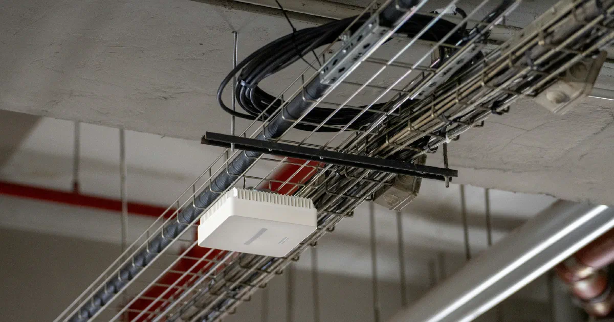 White network box mounted on a metal wire cable tray below a concrete ceiling, with pipes and cables around it