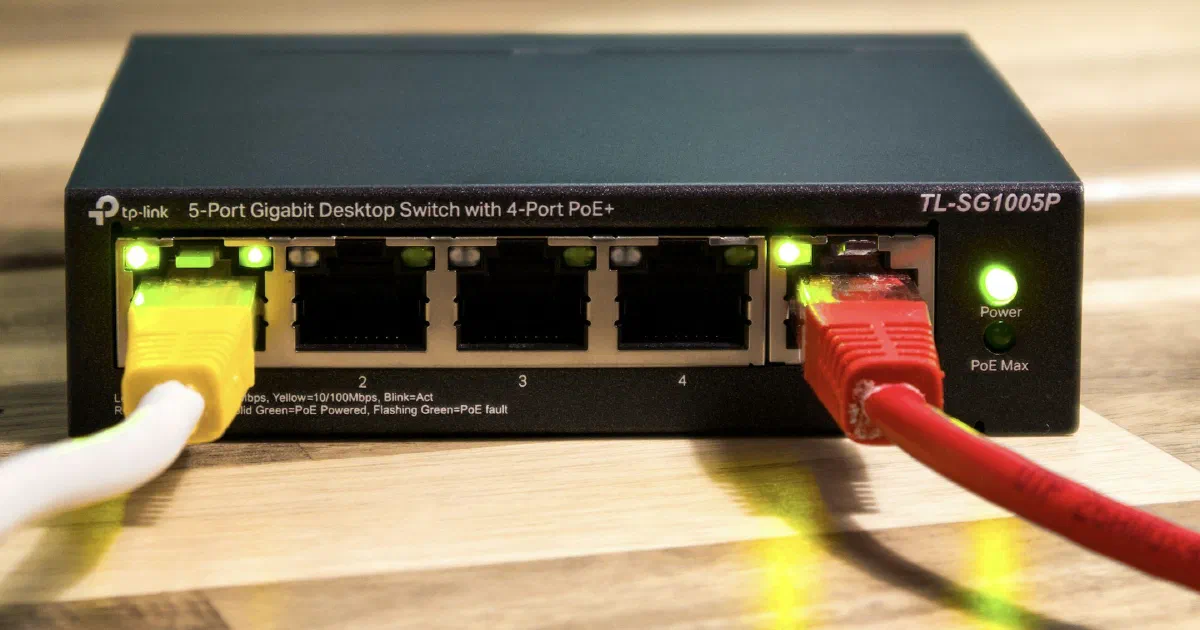 A black TP-Link five-port gigabit desktop switch on a wooden table with a yellow and a red Ethernet cable plugged in and green port lights on