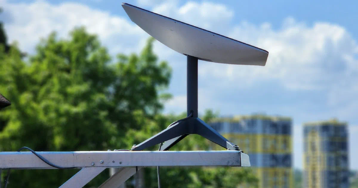 Rectangular satellite internet dish on a black tripod mount fixed to a metal rail, with trees and two apartment towers behind under a blue sky