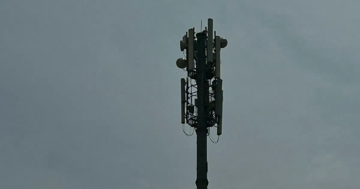 Top of a cellular tower with panel antennas and dish antennas against a gray, overcast sky