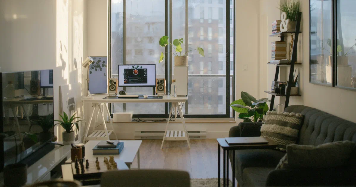 Narrow sunlit apartment living room with a desk and monitor by a tall window, a TV on the left and a gray sofa on the right