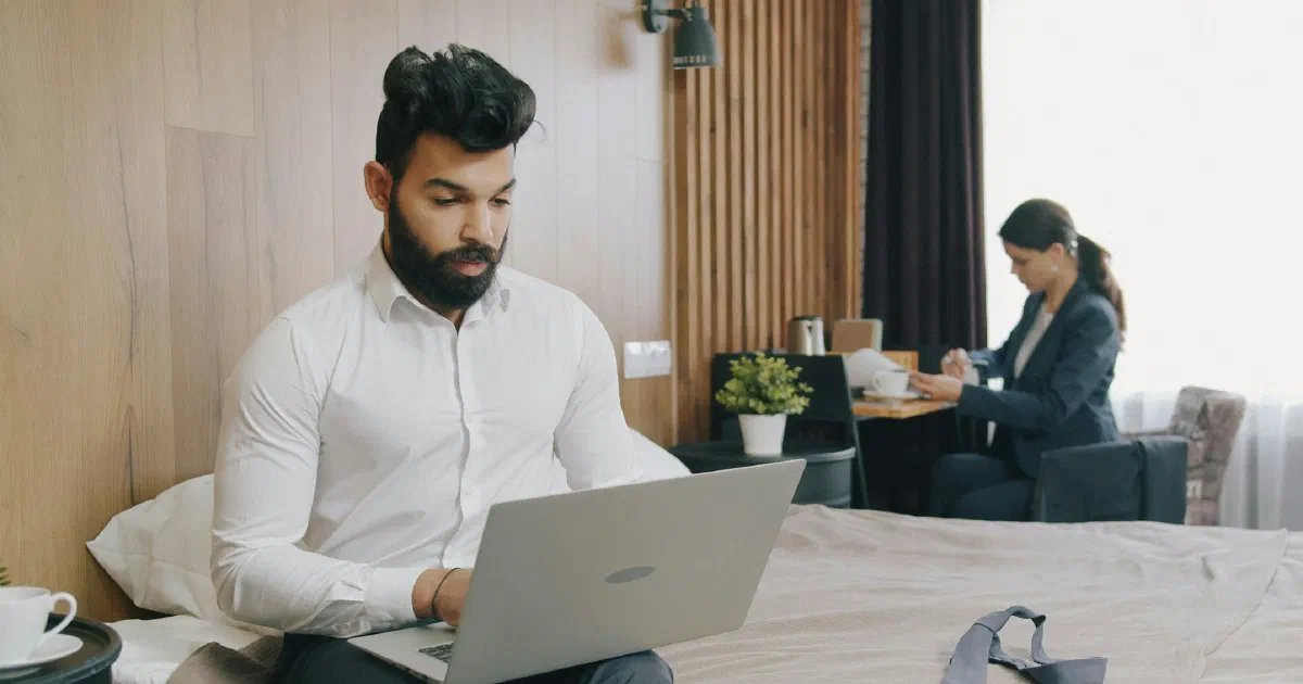 Man working on a laptop while sitting on a hotel bed, with a suitcase at the foot of the bed and a woman writing at a desk behind him