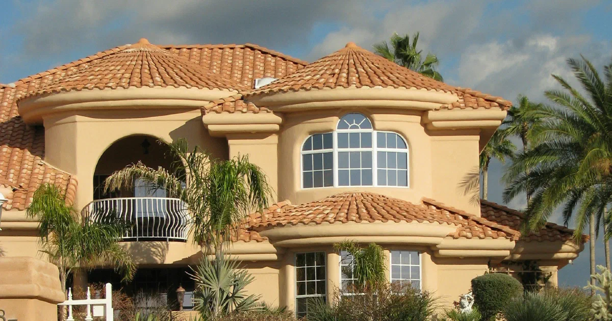 Two-story stucco house with a red tile roof, an arched balcony and palm trees under a cloudy blue sky