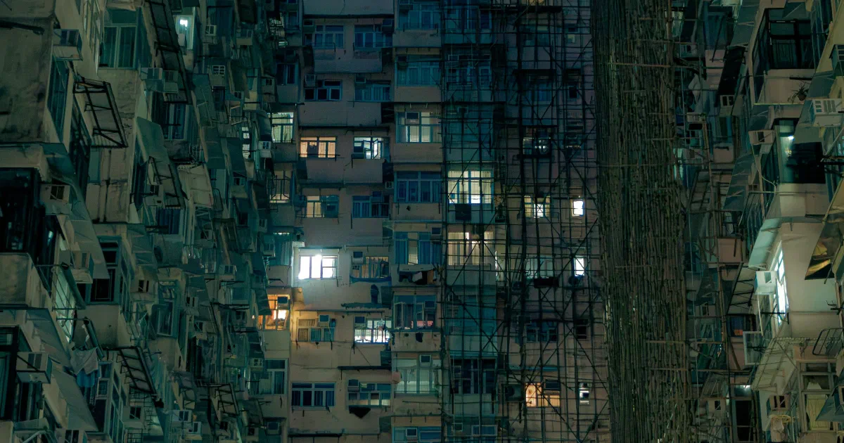Dense apartment blocks at dusk with lit windows, air-conditioning units and bamboo scaffolding