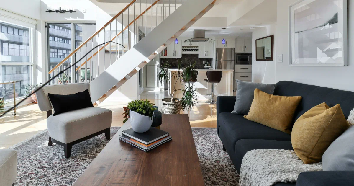 Bright two-level living room with an open staircase, a kitchen behind it, a dark sofa and a wooden coffee table
