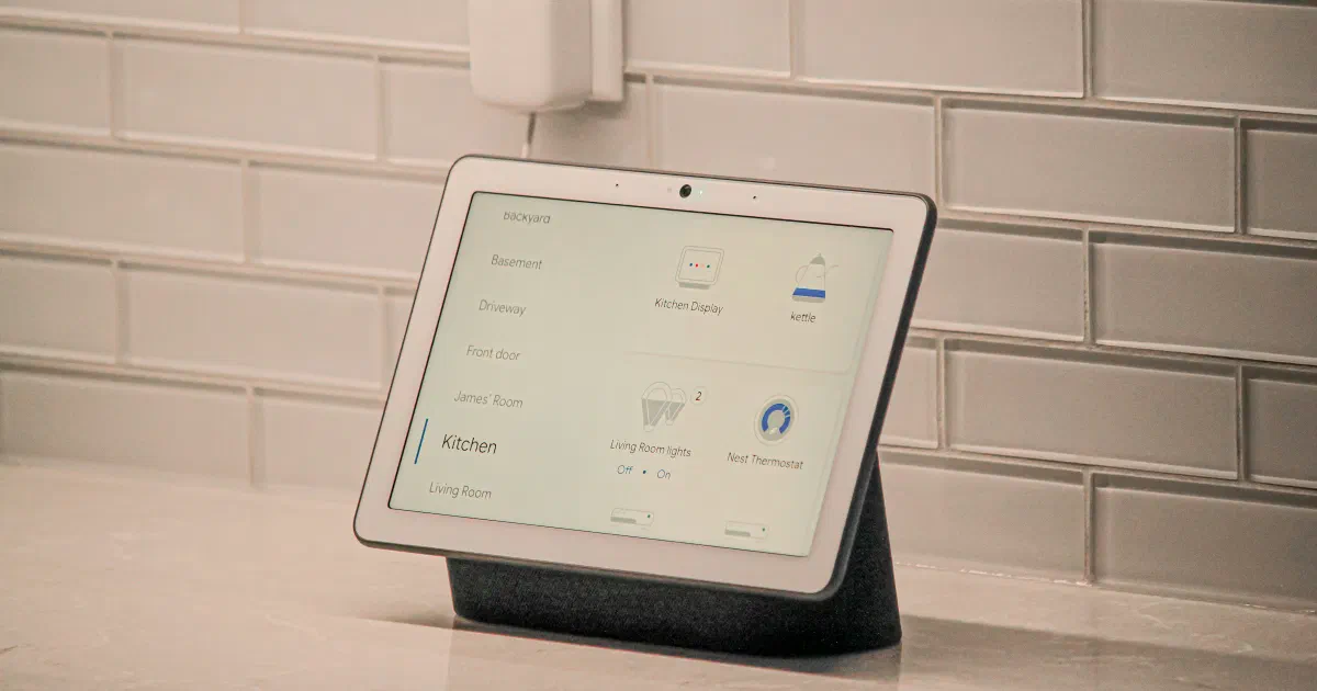 A Google Nest Hub Max smart display on a kitchen counter showing Google Home room and device controls, in front of a white subway-tile backsplash