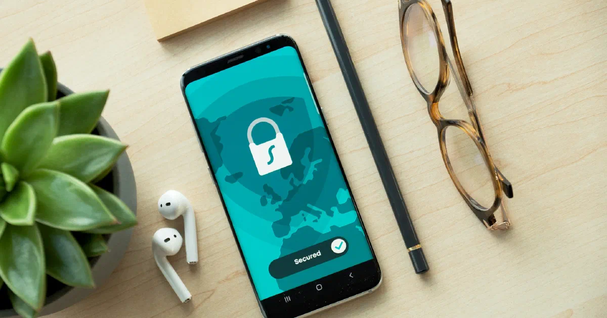Smartphone on a wooden desk showing a padlock icon and a Secured button, beside earbuds, a pencil, glasses and a succulent