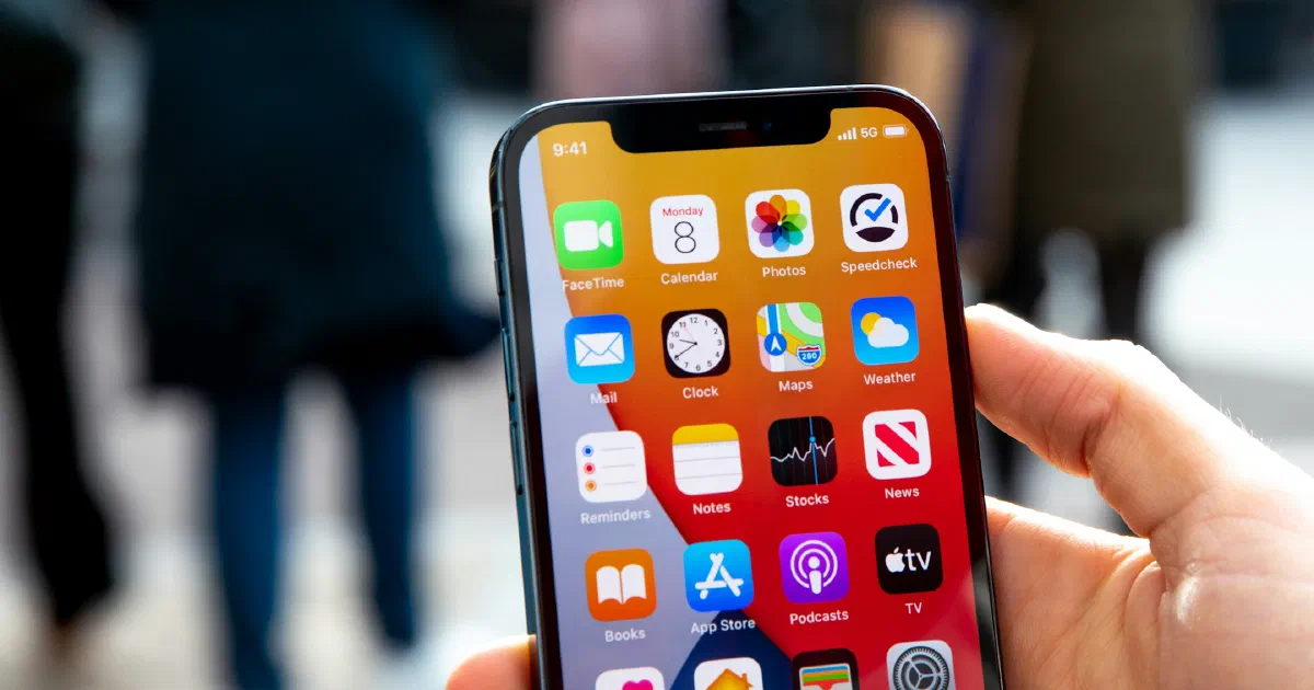 Hand holding an iPhone showing its home screen, with blurred shoppers walking on a city street behind it