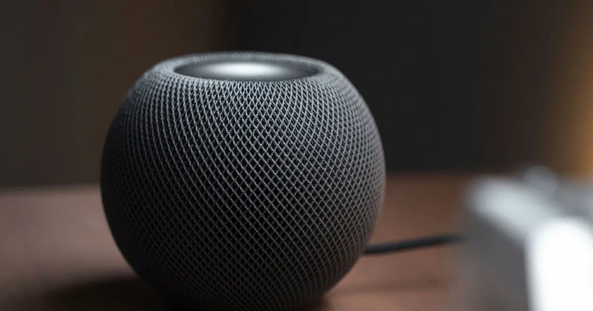 A dark gray HomePod mini smart speaker on a wooden table, with a blurred object in the foreground