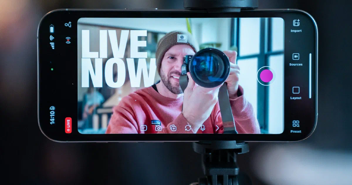 Smartphone on a tripod showing a live-streaming app with a LIVE NOW banner over a man holding a camera