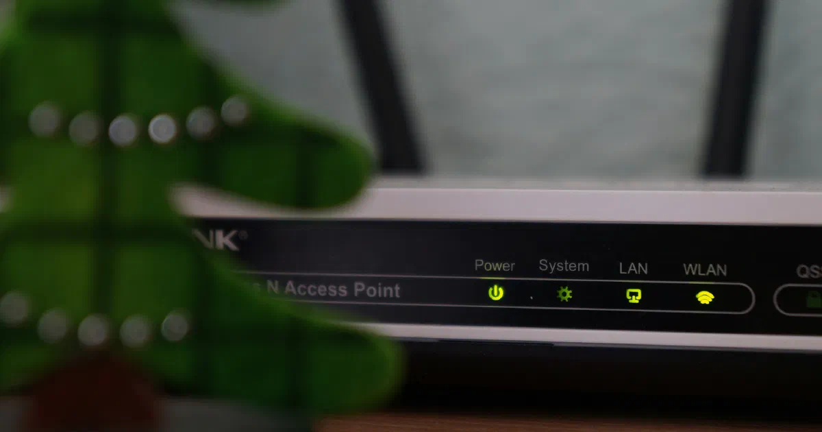 Front of a black wireless router with its Power, System, LAN and WLAN status lights glowing green, behind a blurred felt decoration